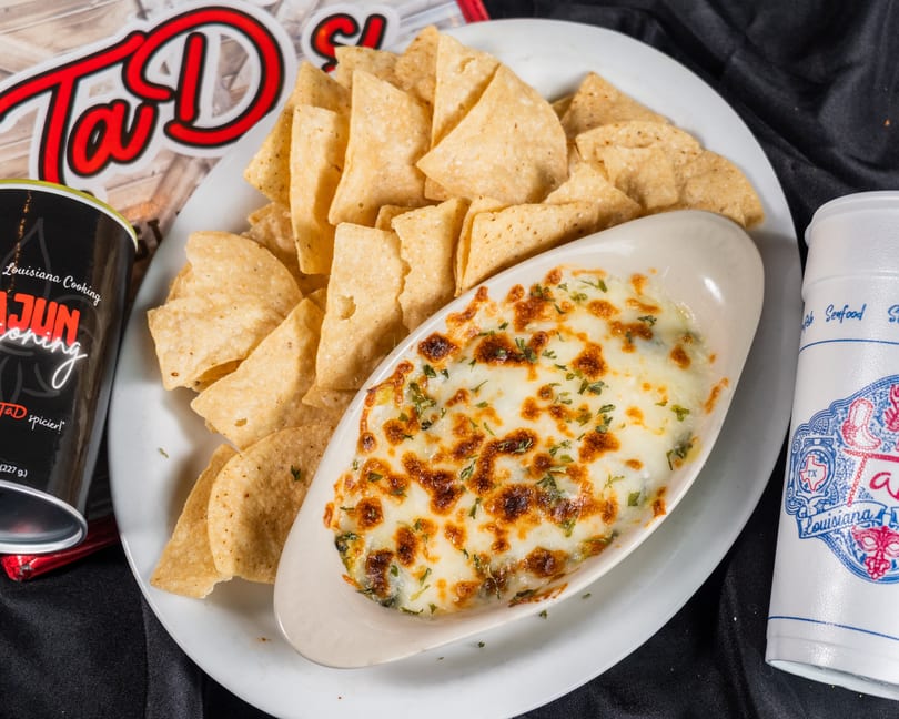 A white plate holds a boat-shaped dish of cheesy dip with tortilla chips, a drink, and seasoning.