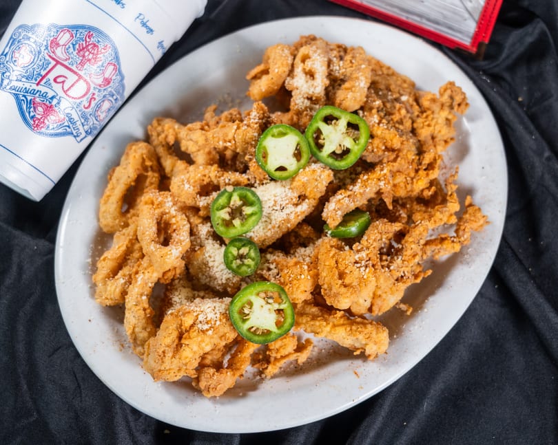 A white plate of golden fried alligator with parmesan cheese and jalapeño slices, next to a Taj's cup.