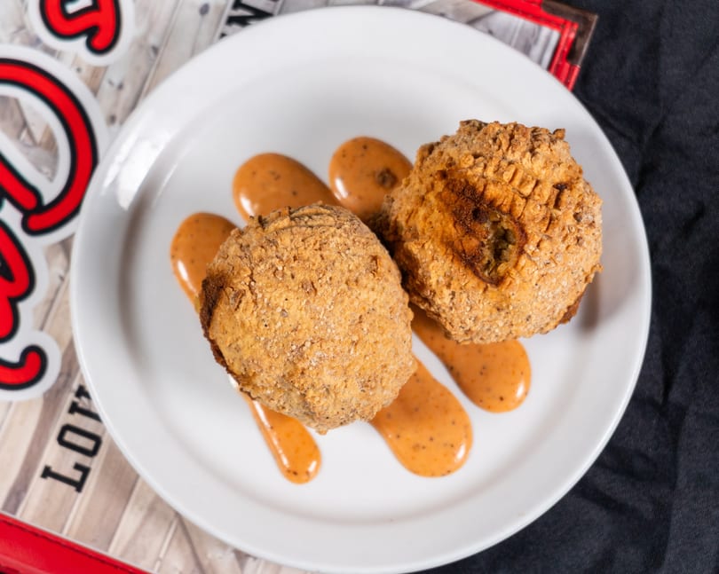 Two golden-brown fried hushpuppies with a creamy orange sauce drizzled on a white plate.
