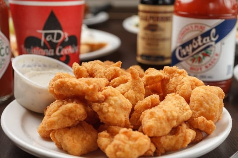 A plate of golden-brown fried popcorn shrimp with dipping sauce, a red cup, and hot sauce bottles.