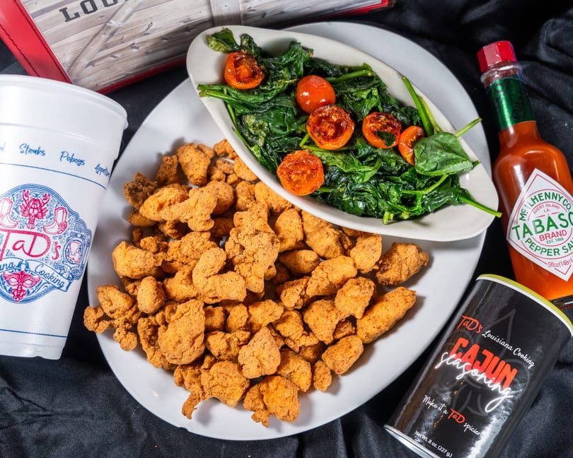 Fried chicken, spinach with tomatoes, a drink, Tabasco, and Cajun seasoning on a black cloth.